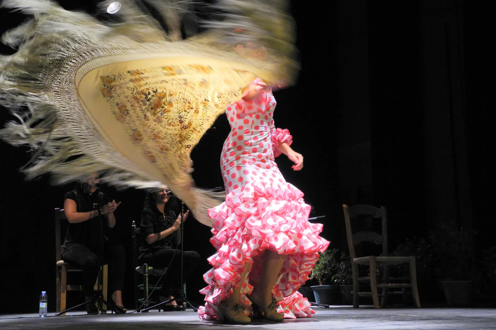 Tanečnice flamenca v růžových puntíkatých šatech, která se točí na tmavém pódiu a kolem ní víří krémový třásnitý šál
